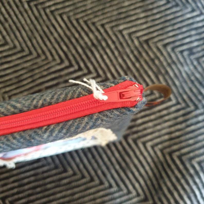 May include: Close-up of a red zipper on a gray and blue tweed fabric pouch. The zipper is partially open and the fabric is textured.