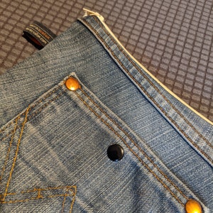May include: A blue denim pouch with a zipper closure and two gold metal rivets. The pouch has a small pocket on the front with a black button.