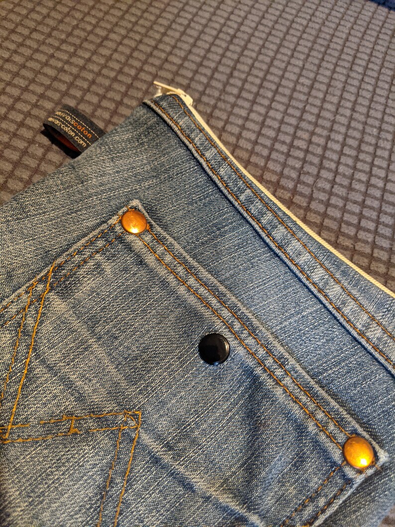 May include: A blue denim pouch with a zipper closure and two gold metal rivets. The pouch has a small pocket on the front with a black button.
