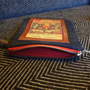 May include: A blue denim zippered pouch with a brown leather patch featuring a vintage poster for a "Shock Show" at the Paramount Theatre. The poster features a skull and crossbones and the text "We Dare to Show Dr. Brundell's Shock Show".