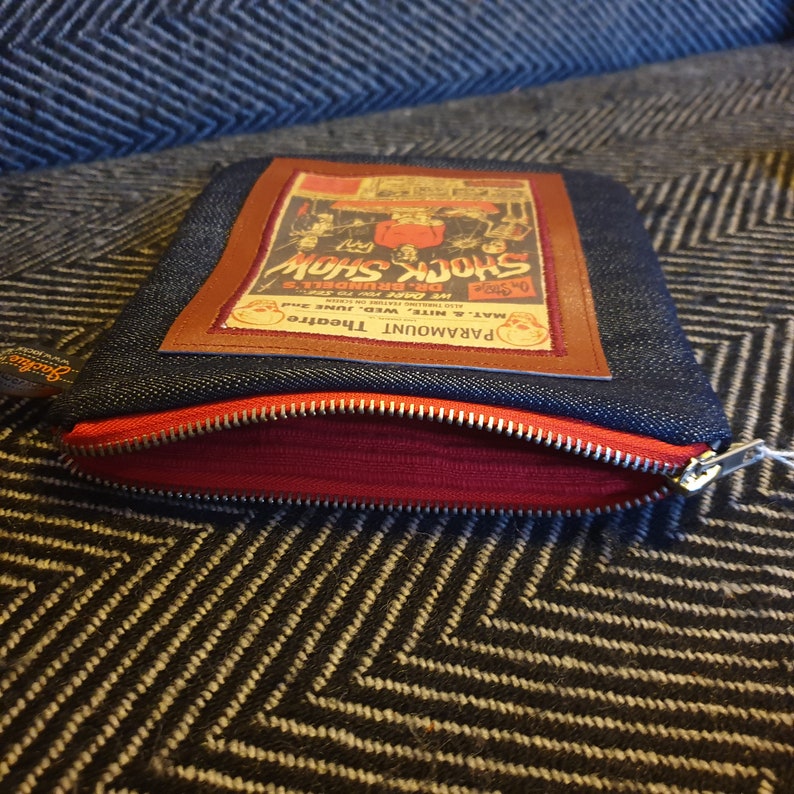 May include: A blue denim zippered pouch with a brown leather patch featuring a vintage poster for a "Shock Show" at the Paramount Theatre. The poster features a skull and crossbones and the text "We Dare to Show Dr. Brundell's Shock Show".