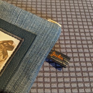 May include: A blue denim zippered pouch with a black leather trim and a small brown and white fabric patch. The pouch has a label that reads "Jackie fileu" and "www.jackie-fileu.com".