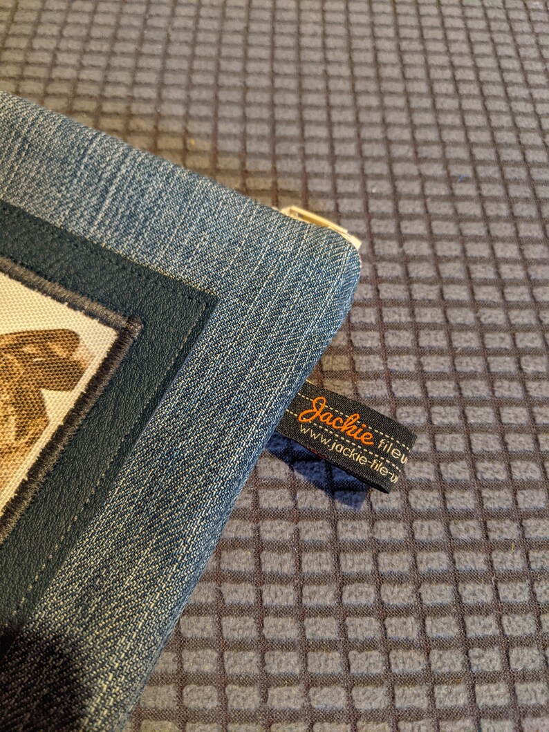 May include: A blue denim zippered pouch with a black leather trim and a small brown and white fabric patch. The pouch has a label that reads "Jackie fileu" and "www.jackie-fileu.com".
