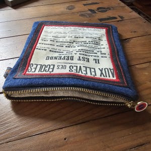 May include: A blue denim pouch with a brass zipper and a rectangular patch featuring French text. The patch is bordered with red and black stitching. The pouch is on a wooden surface.