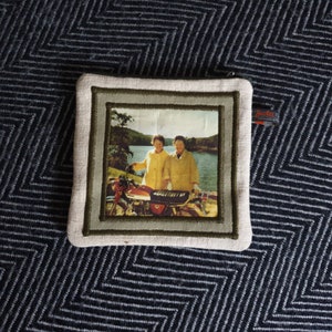 May include: A beige fabric pouch with a green embroidered frame. The frame contains a photo of two people standing next to a motorcycle in front of a lake. The photo is in color and has a vintage feel.