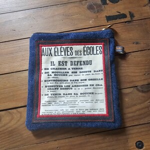 May include: A denim pouch with a rectangular label featuring French text. The label is framed with red and black stitching. The pouch has a zipper closure and a small tag.