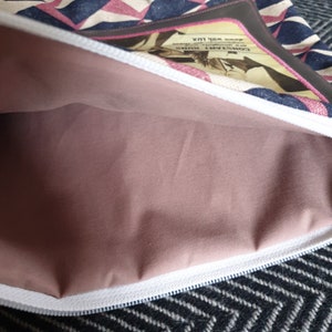 May include: A pink zippered pouch with a white zipper and a black and white geometric pattern on the outside. The pouch is open and the inside is lined with a soft pink fabric. The pouch has a small pocket on the outside with the text "CONSTANT RUNS" and "DOWN WITH LUX" printed on it.