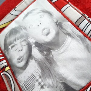 May include: A black and white photograph of two girls pulling funny faces. The girl on the left is wearing glasses and has her mouth open. The girl on the right is sticking her tongue out. The photo is framed in a red and white fabric.