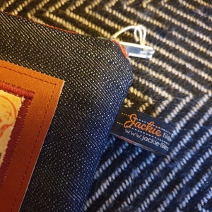 May include: A dark blue denim zippered pouch with a brown leather patch and a black and orange label that reads "Jackie filer www.jackie-filer".