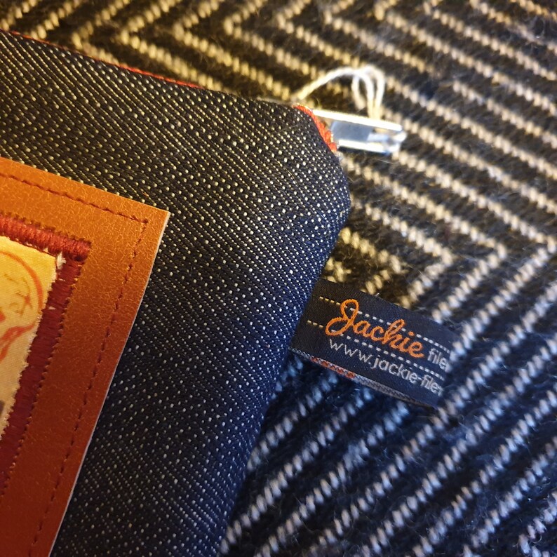 May include: A dark blue denim zippered pouch with a brown leather patch and a black and orange label that reads "Jackie filer www.jackie-filer".