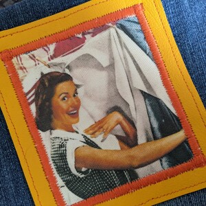 May include: A vintage-style patch with a yellow leather border and an orange stitched inner frame. The patch features a woman smiling and holding a white sheet, with a red and white background.
