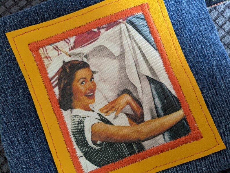 May include: A vintage-style patch with a yellow leather border and an orange stitched inner frame. The patch features a woman smiling and holding a white sheet, with a red and white background.