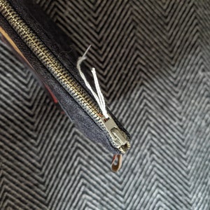 Puede incluir: Una bolsa negra con cremallera con una cremallera dorada y un hilo blanco que cuelga del tirador de la cremallera. La bolsa est&aacute; sobre una tela de patr&oacute;n de chevron gris y blanco.