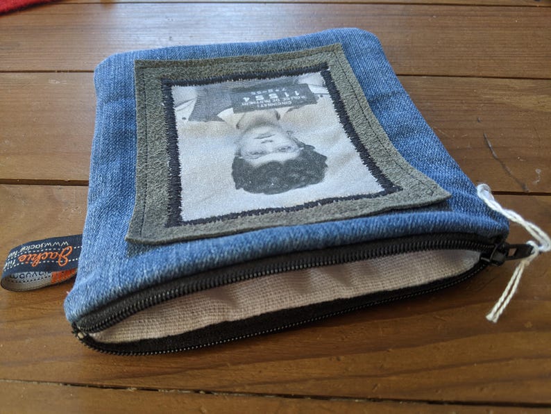 May include: A blue denim zippered pouch with a fabric lining. The pouch features a framed image of a person's face with a mugshot style photo and text that reads "WANTED".