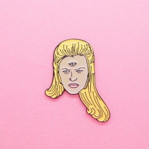 Puede incluir: Un pin de esmalte amarillo con una mujer de dibujos animados con cabello rubio y tres ojos. La mujer tiene una expresión seria.