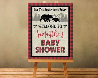 Puede incluir: Un letrero de baby shower sobre un caballete con un borde a cuadros rojo y negro. El letrero dice "Let the Adventure Begin" con siluetas de osos y "Welcome to Samantha's Baby Shower" en texto rojo y negro.