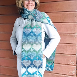 Może przedstawiać: Kobieta w białej kurtce i białych spodniach z niebiesko-zielono-białym dzianym szalikiem z wzorem serca.