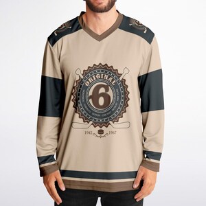 May include: A long-sleeved hockey jersey with a tan background and black and brown stripes. The jersey features a large brown circle with the word "ORIGINAL" and the number "6" inside. The circle is surrounded by the words "MONTREAL, TORONTO, BOSTON, CHICAGO, DETROIT, NEW YORK" and the years "1942" and "1967".
