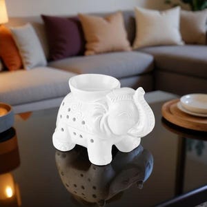 Puede incluir: Un calentador de aceite con forma de elefante de cerámica blanca con un cuenco en la parte posterior. El elefante tiene tallas detalladas y pequeños agujeros para la difusión de la luz y la fragancia. Se encuentra sobre una superficie negra reflectante.