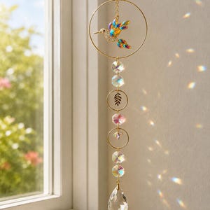 Può includere: Un acchiappasole sospeso con un anello in metallo dorato e un ornamento centrale a forma di colibrì decorato con cristalli iridescenti. La catena verticale include perline di cristallo trasparente sfaccettato, piccoli anelli dorati con motivi a foglia e un grande cristallo a goccia.