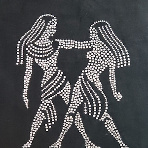 Op de afbeelding: Een zwarte stoffen achtergrond met een ontwerp van het sterrenbeeld Tweelingen in zilveren strass. Het woord "GEMINI" is in strass onder het teken geschreven.