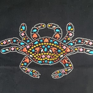 May include: A crab design made of colorful rhinestones on a black background. The crab is decorated with various shapes, including hearts, stars, and diamonds, in shades of orange, pink, blue, and yellow. The rhinestones are outlined with silver beads.