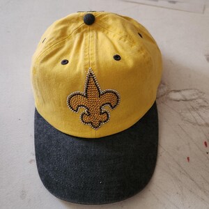 May include: A yellow baseball cap with a black brim. The front of the cap features a fleur-de-lis design made of rhinestones.
