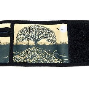 Puede incluir: Una cartera de tela negra con cierre de velcro. La parte delantera de la cartera presenta un fondo beige con una silueta negra de un árbol con raíces y un pájaro volando por encima.