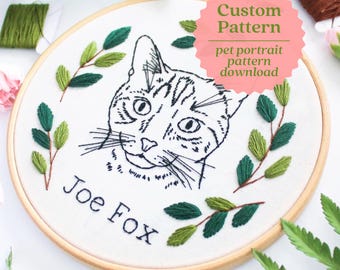 May include: A white embroidery hoop with a black line drawing of a cat's face, surrounded by green embroidered leaves. The hoop has a wooden frame and the text "Joe Fox" is embroidered below the cat's face.