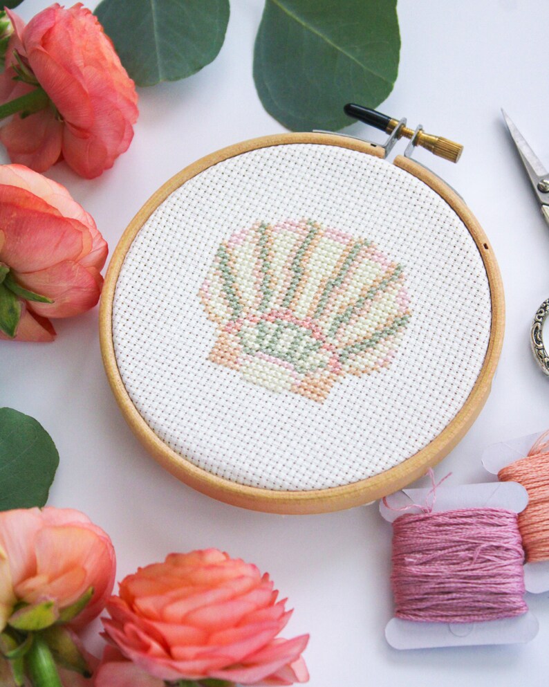 K&ouml;nnte beinhalten: Nahaufnahme eines fertigen Kreuzstich-Stickprojekts in einem h&ouml;lzernen Stickrahmen. Das Design ist eine rosa und gr&uuml;ne Muschel. Zwei Spulen mit rosa Stickgarn sind im Hintergrund sichtbar.