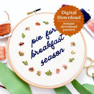 May include: Embroidered hoop with the text "pie for breakfast season" in navy blue thread. The hoop is surrounded by small embroidered flowers and leaves in shades of pink, green, and brown.