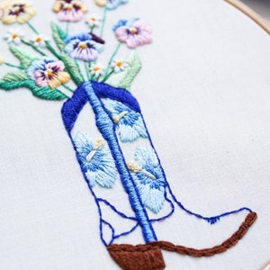 May include: Embroidered hoop art featuring a bouquet of colorful pansies in a blue cowboy boot. The boot is decorated with blue and white flowers.
