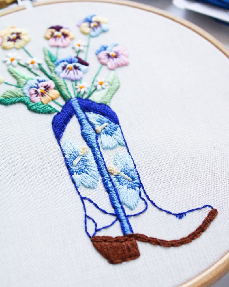 May include: Embroidered hoop art featuring a bouquet of colorful pansies in a blue cowboy boot. The boot is decorated with blue and white flowers.
