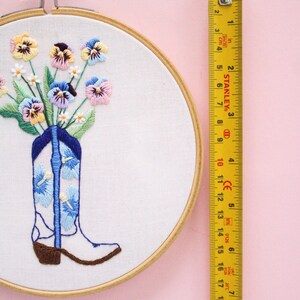 May include: A white cowboy boot with blue and white floral embroidery is filled with a bouquet of colorful pansies and small white flowers. The embroidery hoop is made of wood and has a diameter of 8 inches.