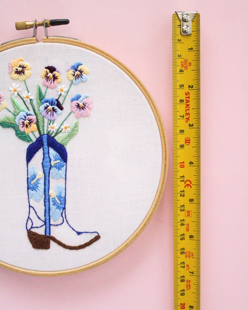 May include: A white cowboy boot with blue and white floral embroidery is filled with a bouquet of colorful pansies and small white flowers. The embroidery hoop is made of wood and has a diameter of 8 inches.