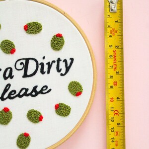 May include: A white embroidery hoop with the phrase "a Dirty lease" stitched in black thread. The hoop is decorated with green and red embroidered olives. A yellow measuring tape is visible to the right of the hoop.