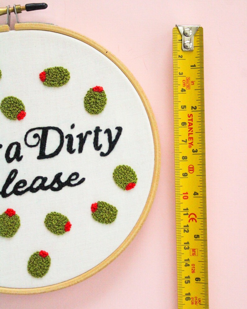 May include: A white embroidery hoop with the phrase "a Dirty lease" stitched in black thread. The hoop is decorated with green and red embroidered olives. A yellow measuring tape is visible to the right of the hoop.