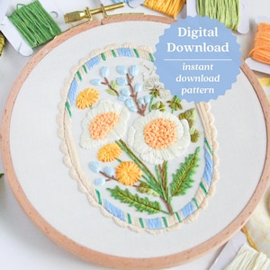May include: An embroidery hoop with a floral design. The design features white flowers with orange centers, yellow accents, and green leaves, all within a blue and cream border. The text "Digital Download" is visible.