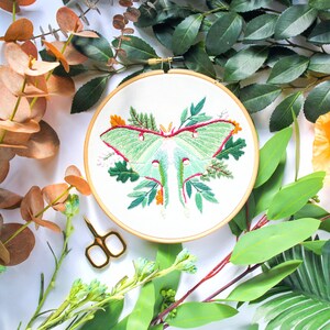 May include: A green and white embroidered moth with red accents on a white fabric background in a wooden embroidery hoop. The hoop is surrounded by green leaves.