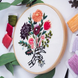 Puede incluir: Arte de bordado en aro que presenta una mano de esqueleto sosteniendo un ramo de flores coloridas, incluyendo rosa, rojo, naranja, morado y verde.