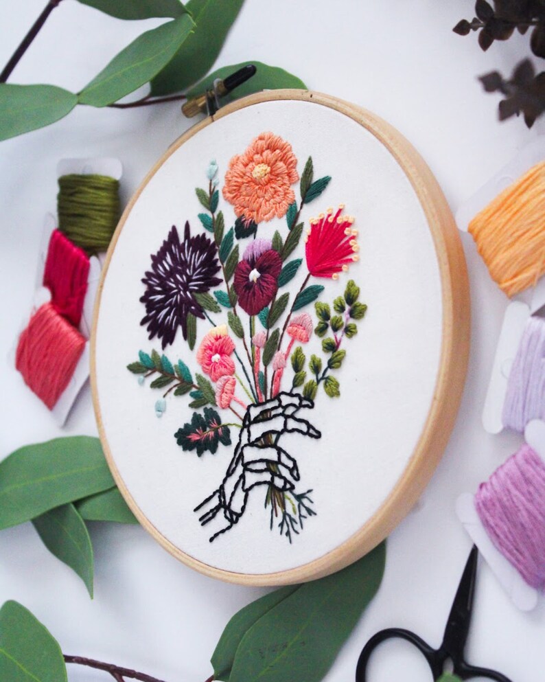 Puede incluir: Arte de bordado en aro que presenta una mano de esqueleto sosteniendo un ramo de flores coloridas, incluyendo rosa, rojo, naranja, morado y verde.