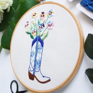 May include: Embroidered hoop art featuring a white cowboy boot with blue and white floral pattern, filled with a bouquet of pink, purple, and yellow pansies.