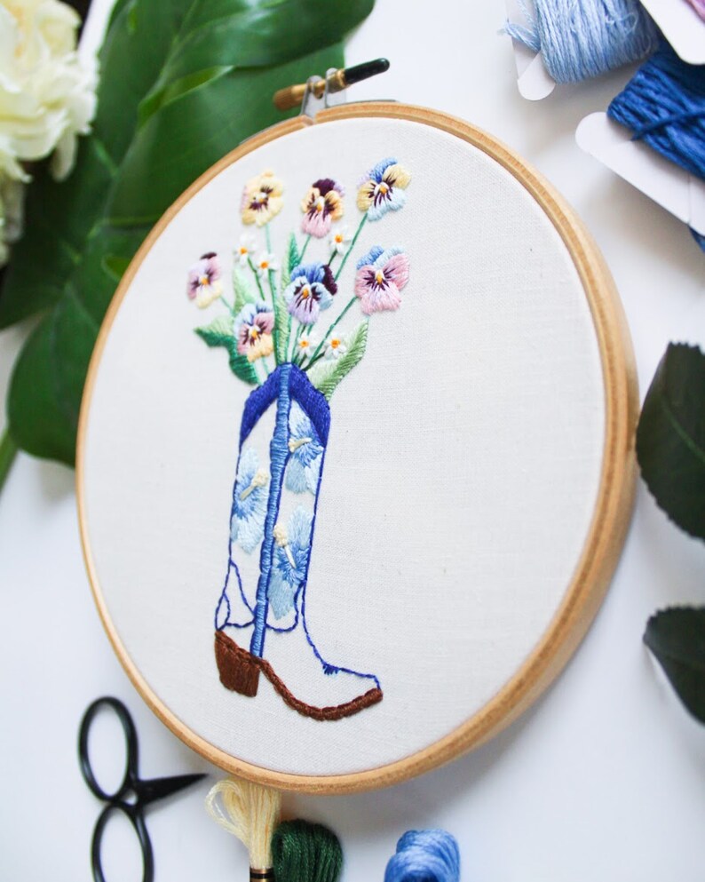 May include: Embroidered hoop art featuring a white cowboy boot with blue and white floral pattern, filled with a bouquet of pink, purple, and yellow pansies.
