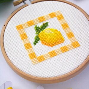 May include: A cross-stitch embroidery hoop featuring a yellow lemon design with green leaves. The lemon is inside a square border with a yellow and orange checkered pattern. The embroidery is set in a wooden hoop.