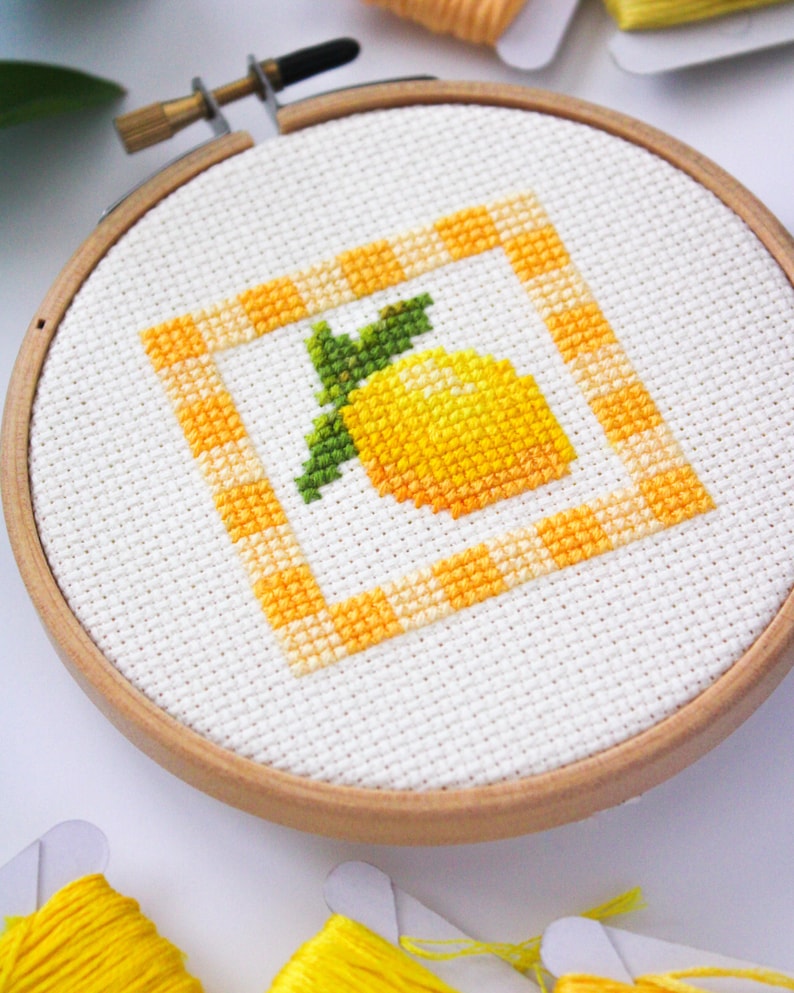 May include: A cross-stitch embroidery hoop featuring a yellow lemon design with green leaves. The lemon is inside a square border with a yellow and orange checkered pattern. The embroidery is set in a wooden hoop.