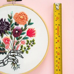 Puede incluir: Arte de bordado en aro que presenta una mano de esqueleto sosteniendo un ramo de flores coloridas. Las flores son rosas, rojas, naranjas, moradas y verdes. El aro tiene un di&aacute;metro de 15 cm.