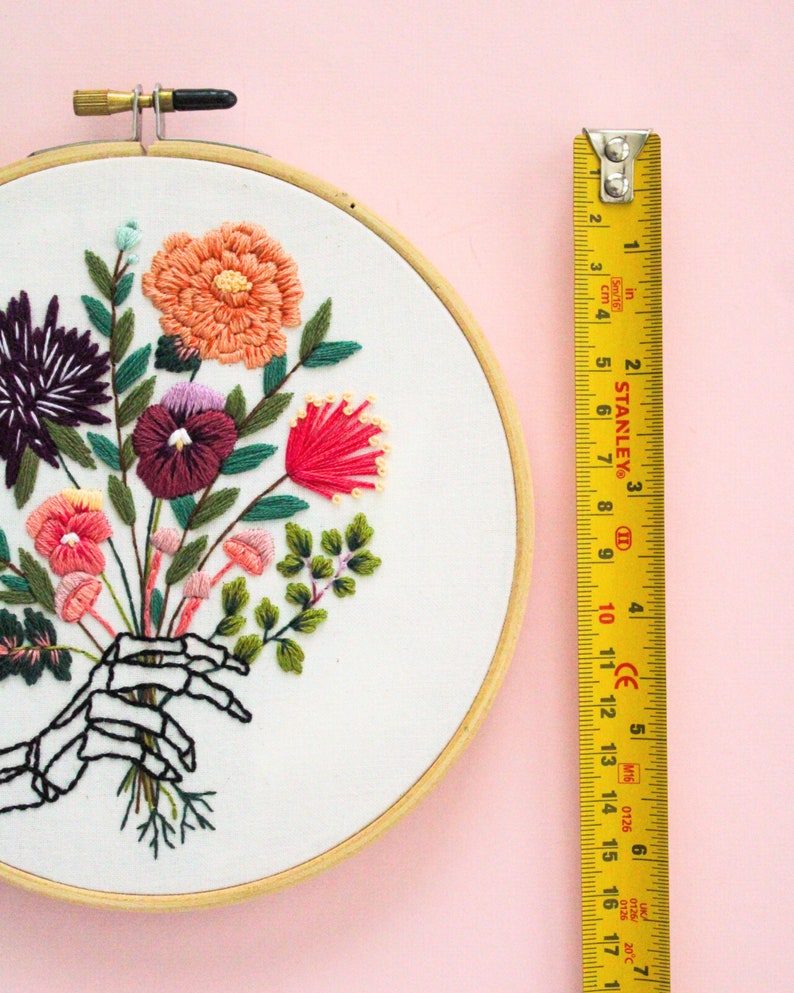 Puede incluir: Arte de bordado en aro que presenta una mano de esqueleto sosteniendo un ramo de flores coloridas. Las flores son rosas, rojas, naranjas, moradas y verdes. El aro tiene un di&aacute;metro de 15 cm.