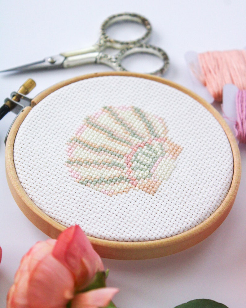 K&ouml;nnte beinhalten: Nahaufnahme eines fertigen Kreuzstich-Stickprojekts in einem Holzreifen. Das Design ist eine pastellrosa, gr&uuml;ne und wei&szlig;e Muschel.