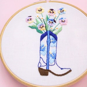 May include: Embroidered hoop art featuring a white cowboy boot with blue and white floral embroidery. The boot is filled with a bouquet of colorful pansies.