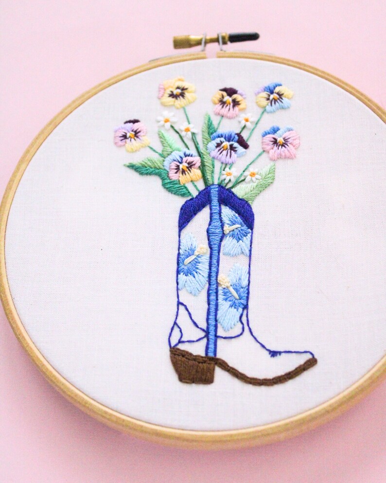 May include: Embroidered hoop art featuring a white cowboy boot with blue and white floral embroidery. The boot is filled with a bouquet of colorful pansies.